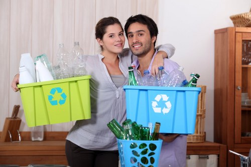 A young couple standing indoors in a room with wooden furniture and neutral-colored curtains, holding three containers filled with empty glass bottles and recyclable plastics. The woman on the left has dark hair tied back and is wearing a light gray cardigan over a white top, holding a green recycling bin with a blue recycling symbol, which contains clear plastic bottles. The man on the right has dark, slightly wavy hair and is wearing a light purple shirt, holding a blue bin with the same recycling symbol, filled with empty glass bottles of various green, clear, and brown tones. In front of him is a smaller light blue container with a dark green recycling symbol, also filled with glass bottles. The containers are positioned in front of a wooden sideboard, and the background shows a casual, domestic setting. The image emphasizes waste separation and recycling, aligning with rubbish removal services provided by Garden Clearance Havering in the Havering area.