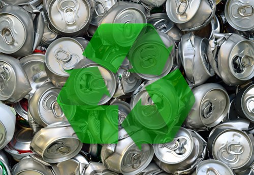A close-up view of numerous discarded aluminium beverage cans piled together, with visible pop tab openings and smooth, metallic surfaces in silver and grey tones. The cans are crushed and stacked irregularly, filling the foreground and extending into the background, creating a dense collection of recyclables. The metallic texture appears reflective, with light glinting off some surfaces, and the cans show slight dents and creases characteristic of used packaging. The scene is set outdoors or in a warehouse environment, with no other objects or background details visible besides the cans. The image features a partially transparent green recycling symbol overlay, indicating waste management or recycling activity, which subtly aligns with rubbish removal services like those offered by Garden Clearance Havering in Essex. Overall, the image highlights metal waste collection, essential to efficient rubbish disposal and recycling efforts relevant to Havering's local waste management services.