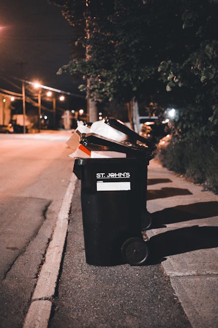 A black wheeled rubbish bin labeled 'ST. JOHN'S' positioned on the pavement next to a curb in a residential area during nighttime. The bin is open and filled with assorted waste, including cardboard b