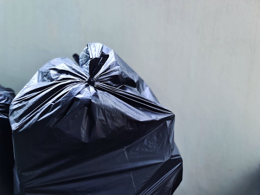 A black wheeled rubbish bin labeled 'ST. JOHN'S' positioned on the pavement next to a curb in a residential area during nighttime. The bin is open and filled with assorted waste, including cardboard b