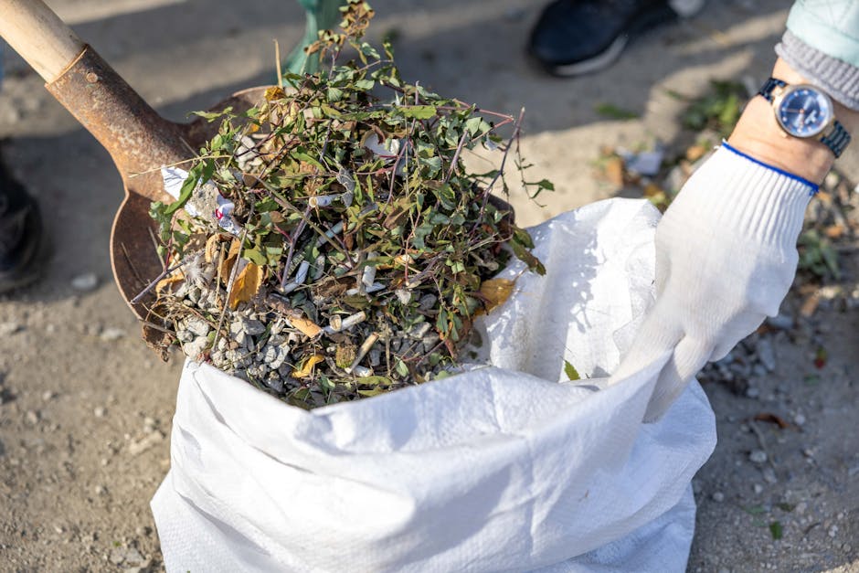 A person wearing a white glove with blue trim, a wristwatch, and light-colored clothing is holding open a large white polypropylene sack filled with garden debris. Inside the sack are a mixture of sma