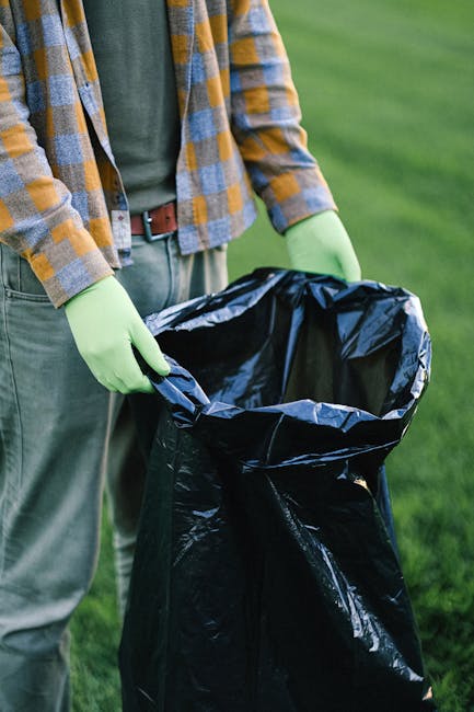 A person wearing a white glove with blue trim, a wristwatch, and light-colored clothing is holding open a large white polypropylene sack filled with garden debris. Inside the sack are a mixture of sma