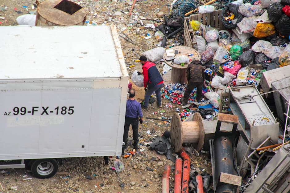 A pile of mixed rubbish and waste materials scattered on an asphalt surface in an outdoor setting, including black and clear plastic rubbish bags, discarded cardboard and paper, a yellow plastic conta